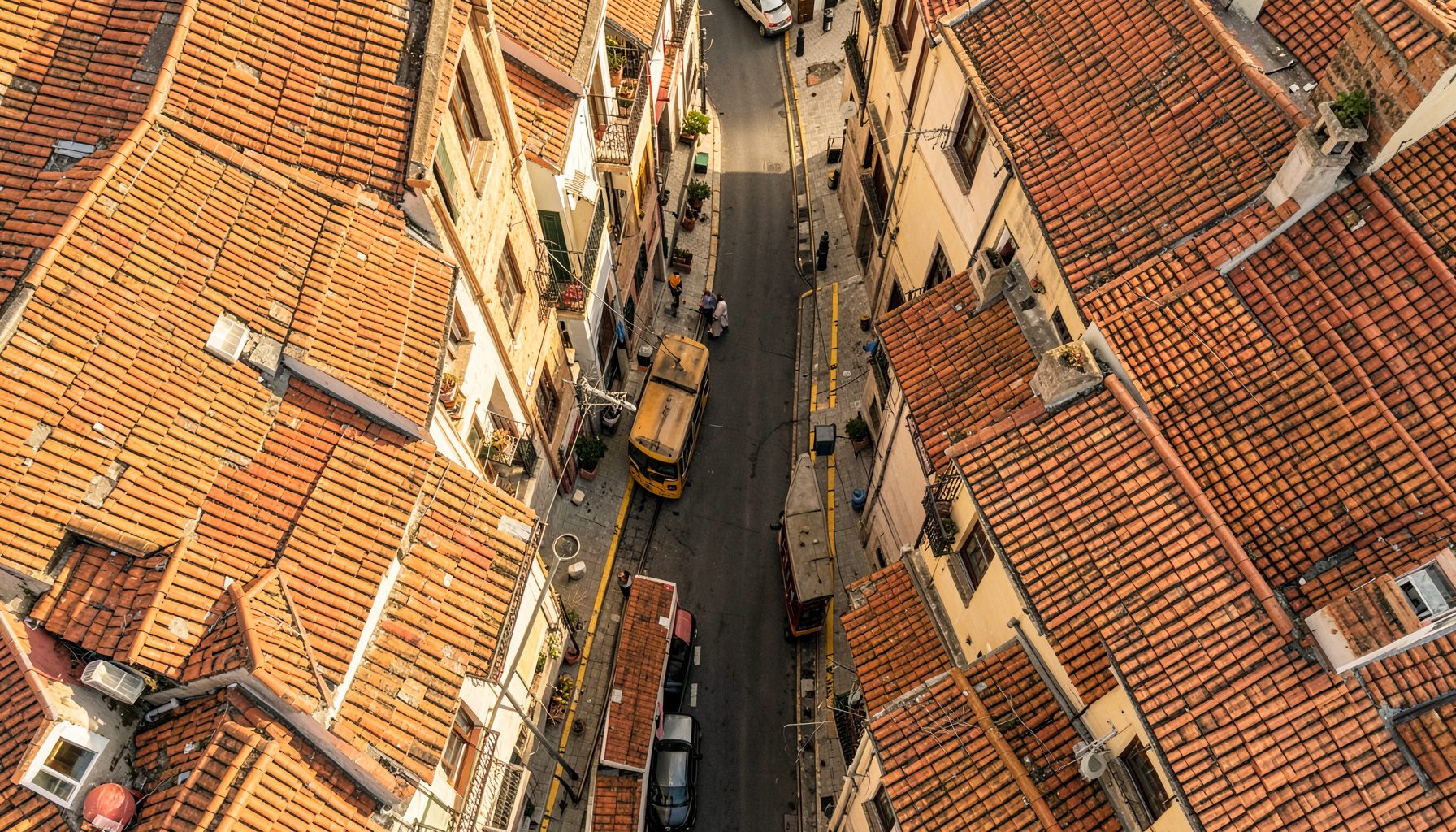 Overhead flat-lay photograph of a sunlit Lisbon street scene with terracotta rooftops and yellow tram tracks viewed from above, warm golden light and soft shadows.